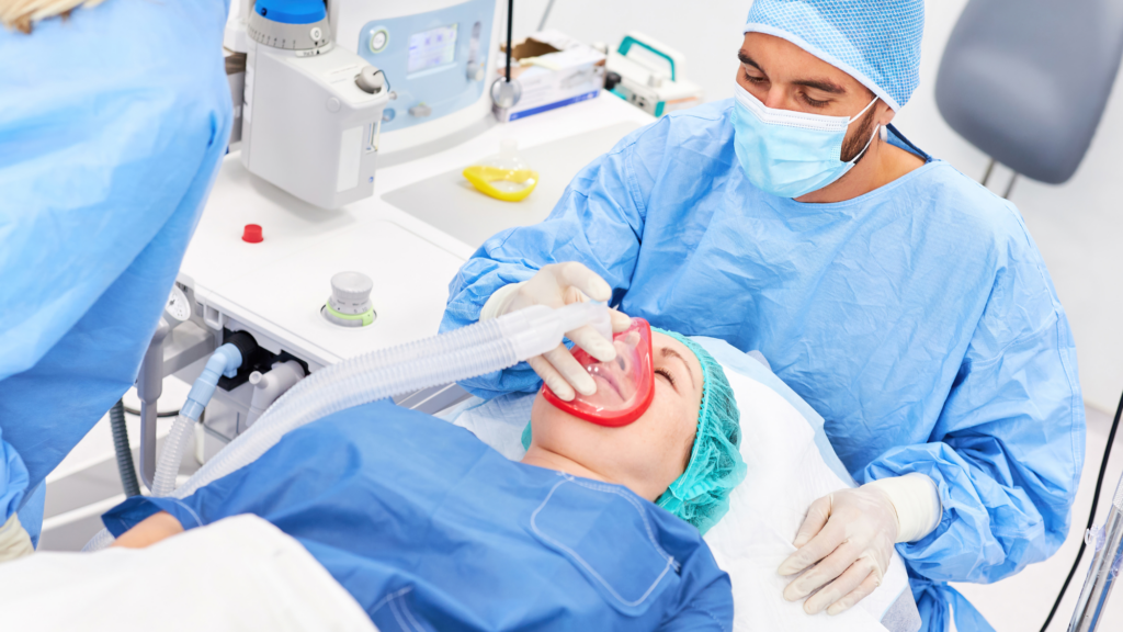 Pasient får narkosebehandling hos tannlegen, med helsepersonell i verneutstyr som overvåker prosedyren.






